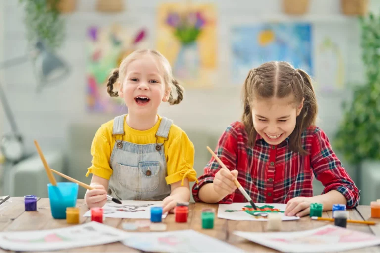 Twee jonge kinderen schilderen en lachen samen aan een tafel in een kleurrijk klaslokaal